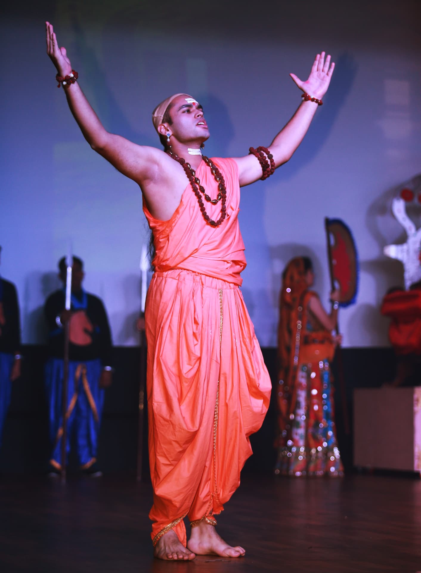 Actor in orange robes with arms outstretched on stage