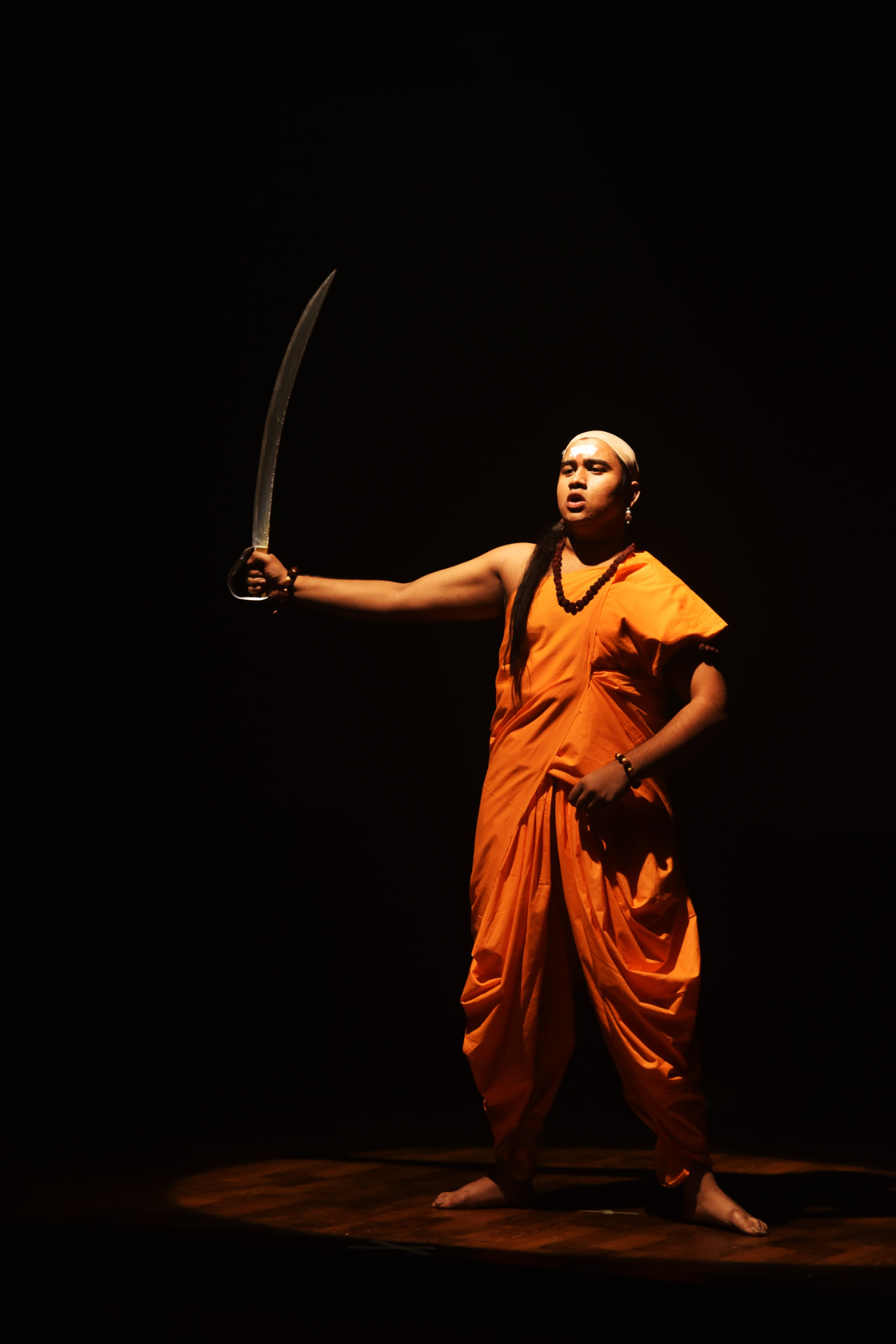 Actor in orange robes holding a curved sword in a spotlight